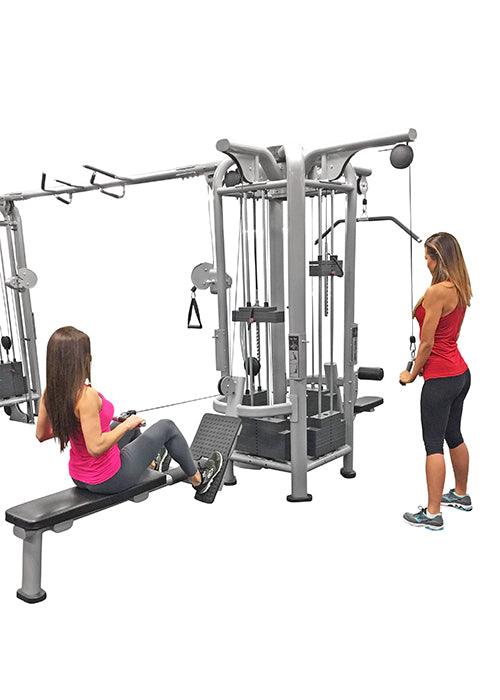 Two women using a gray multi-station gym machine with various cable attachments and weight stacks.