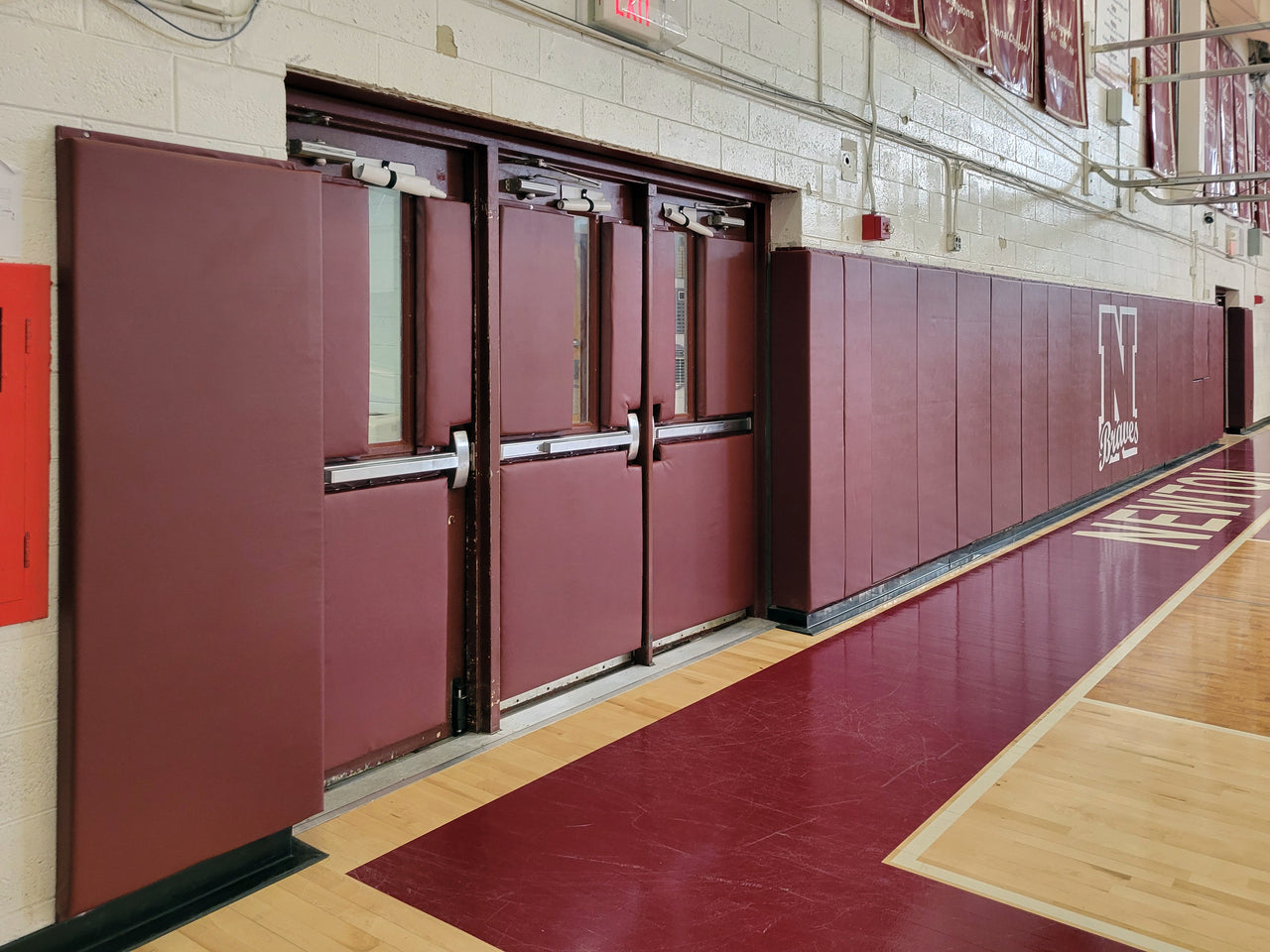 Maroon gym wall padding and padded double doors with a white N Braves logo along a basketball court