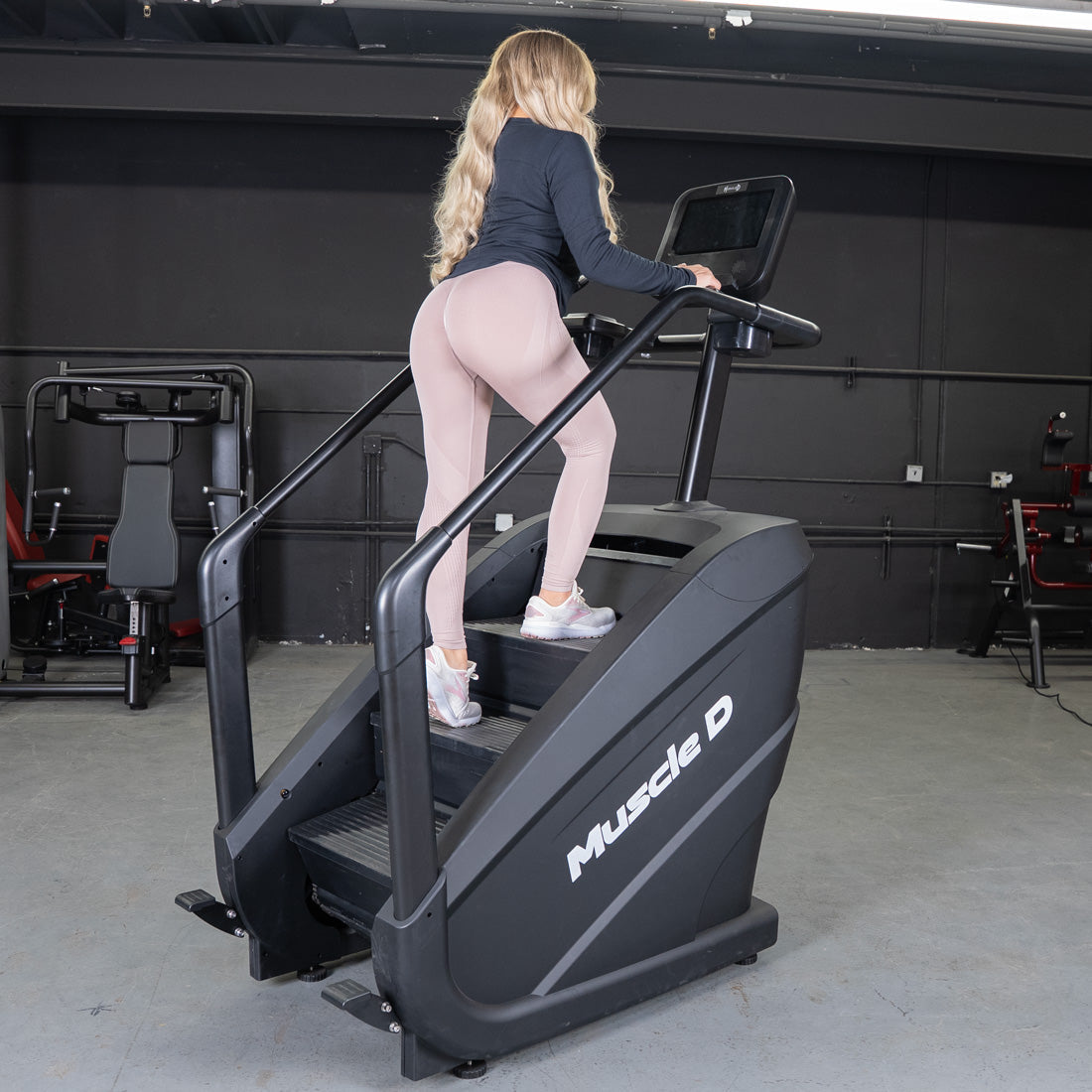 Woman in athletic wear using a black Muscle D Fitness stair climber with a touchscreen in a gym.