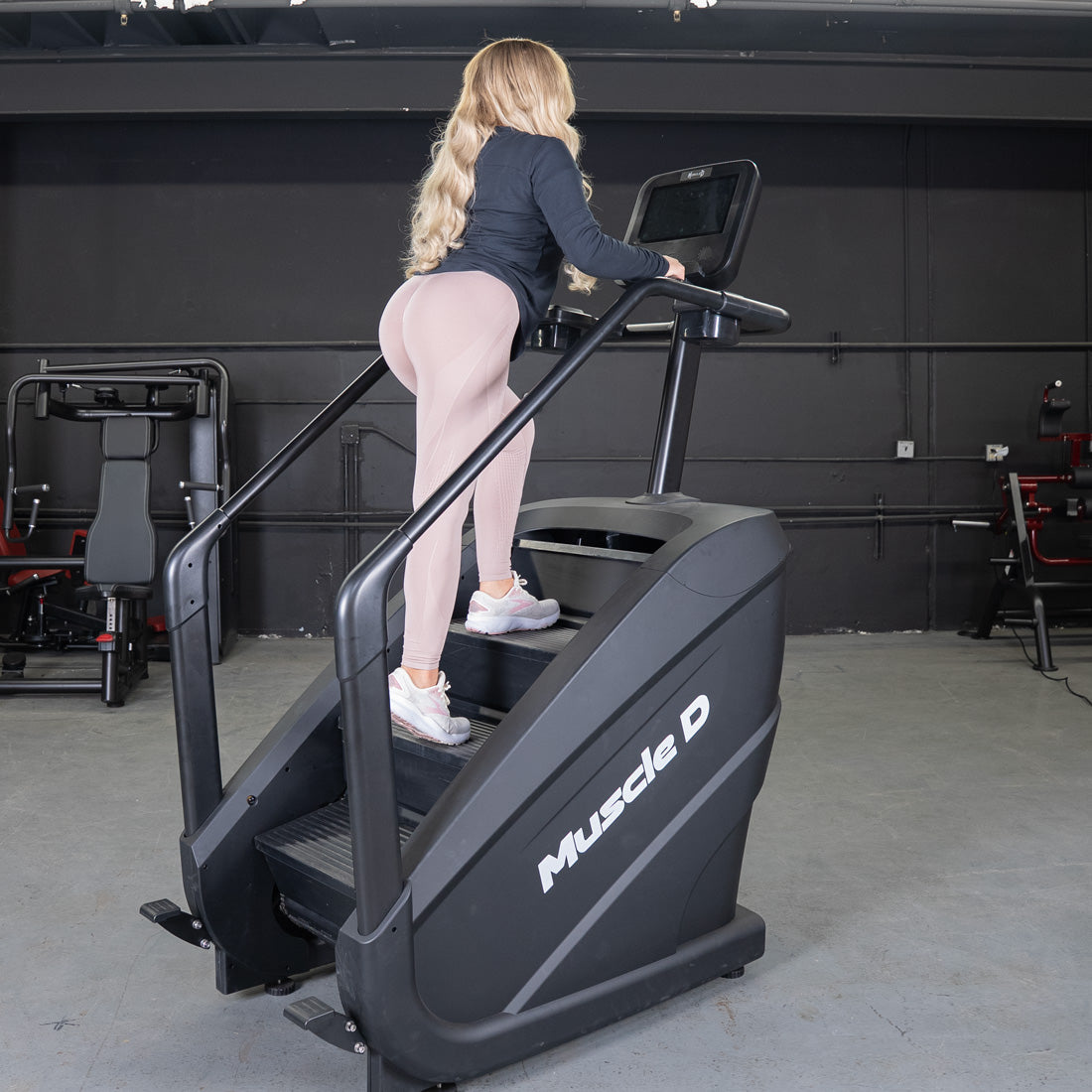 Woman in pink leggings using a black Muscle D Fitness stair climber with a touchscreen display.