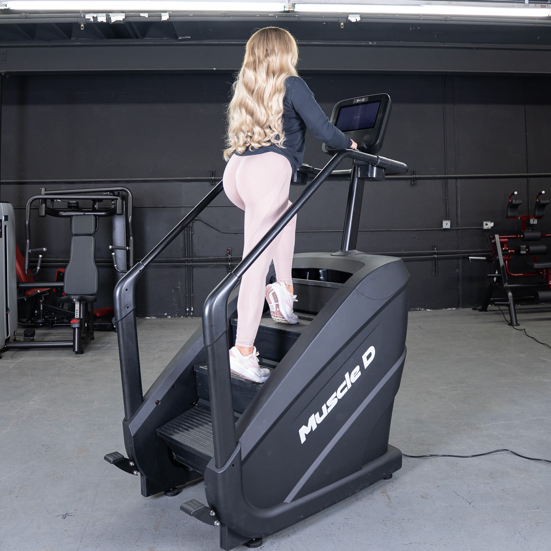 Woman in pink leggings using a black Muscle D Fitness stair climber with a touchscreen in a gym.