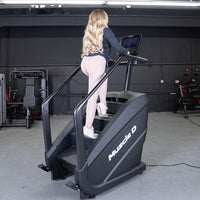Thumbnail for Woman in pink leggings using a black Muscle D Fitness stair climber with a touchscreen in a gym.