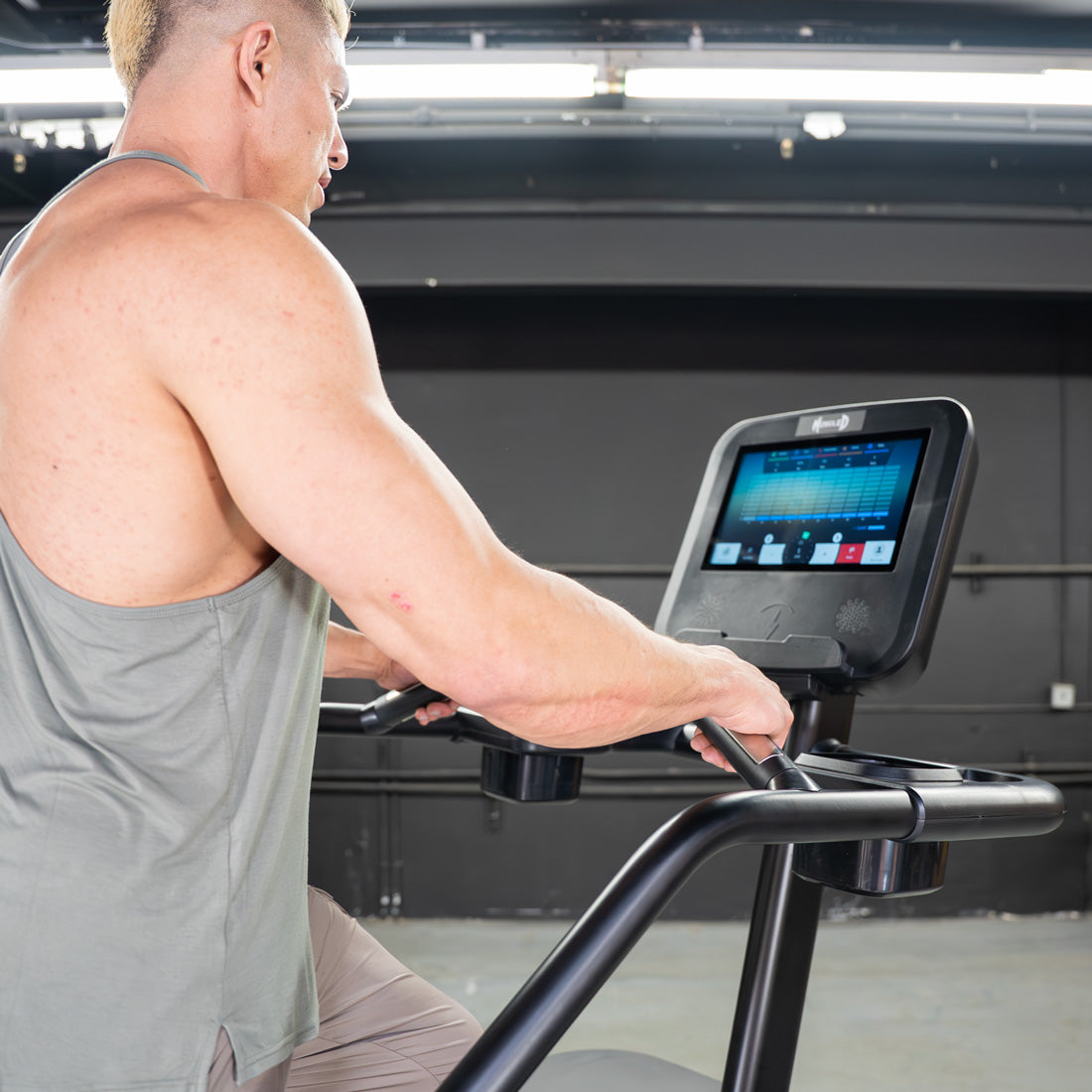 Man in a gray tank top using a black stair climber with a large touchscreen display.