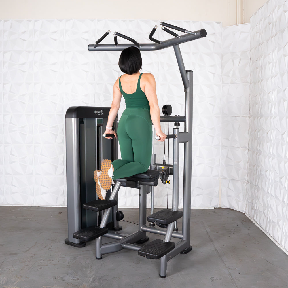 Woman in green using an assisted chin/dip machine, kneeling on the pad and holding dip handles.