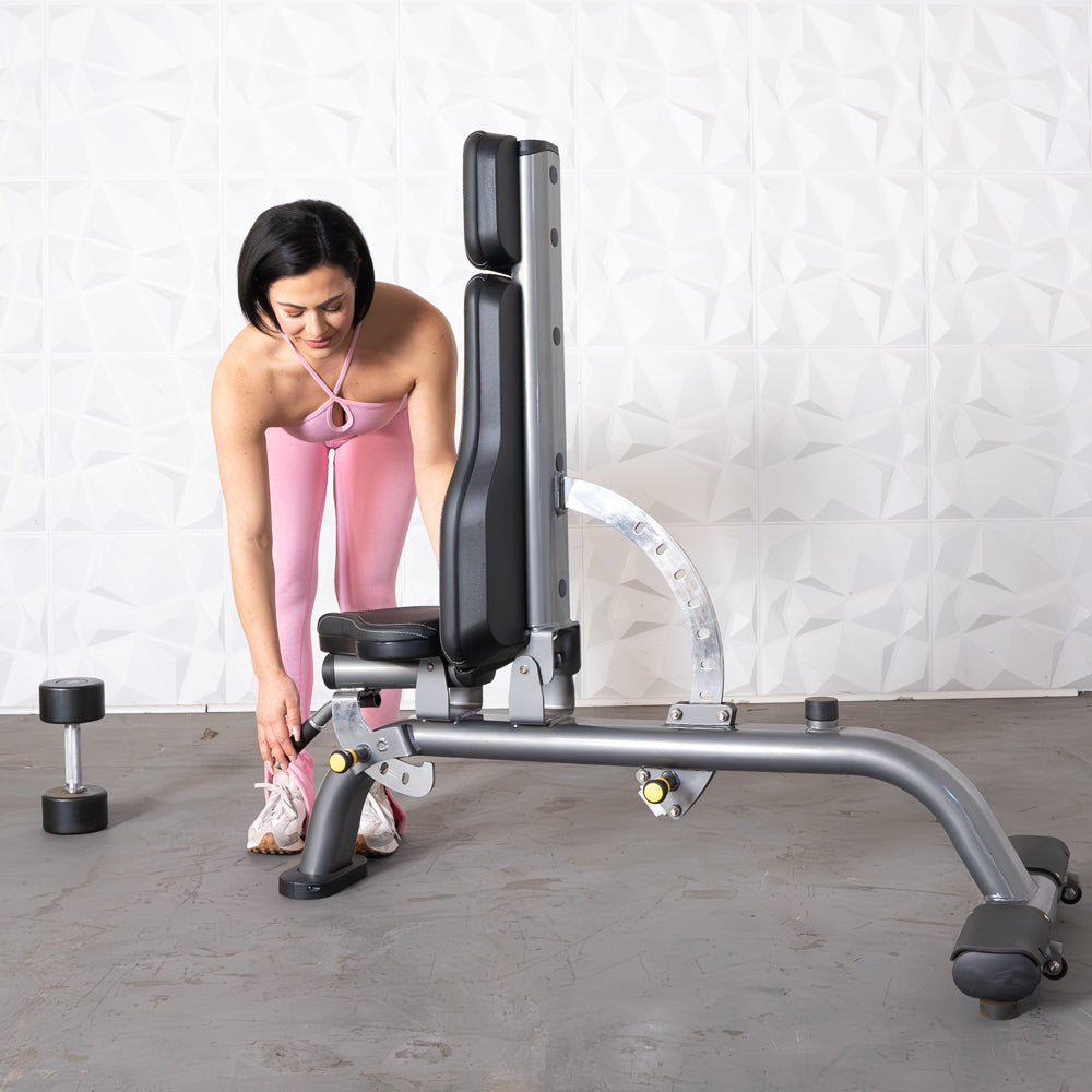 Woman in pink adjusting a gray adjustable weight bench, with a dumbbell on the floor.