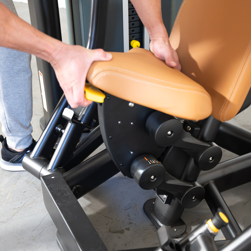 Hands adjusting the tan padded seat and yellow lever of a black commercial chest press machine.