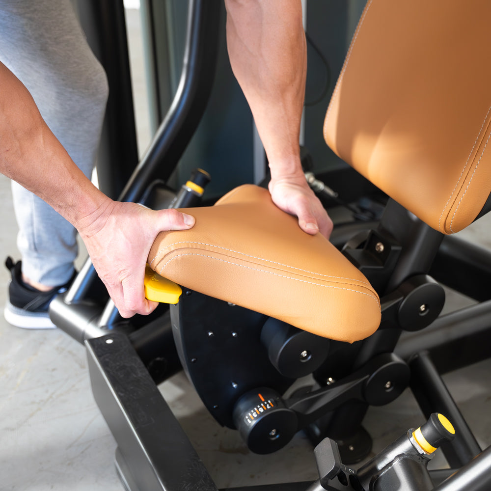Hands adjusting the tan seat pad of a black chest press machine with yellow levers.