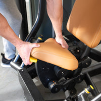 Thumbnail for Hands adjusting the tan seat pad of a black chest press machine with yellow levers.