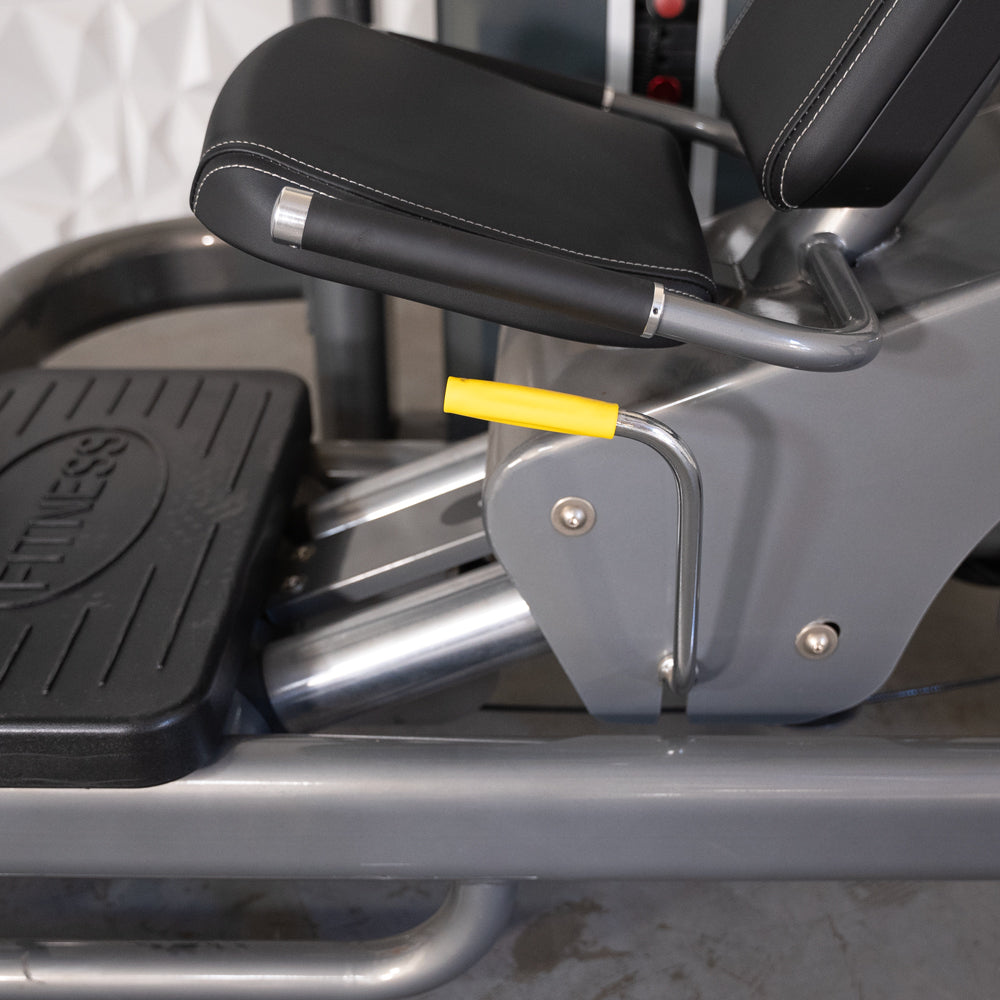Close-up of a leg press machine with black padded seat, yellow adjustment handle, and "FITNESS" on a footplate.