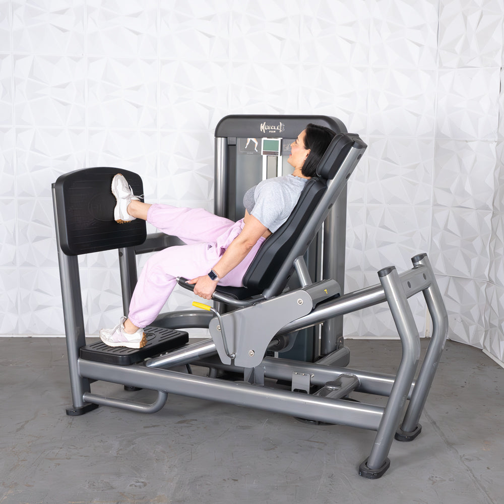 Woman using a gray leg press machine with black pads, wearing pink pants and white sneakers.