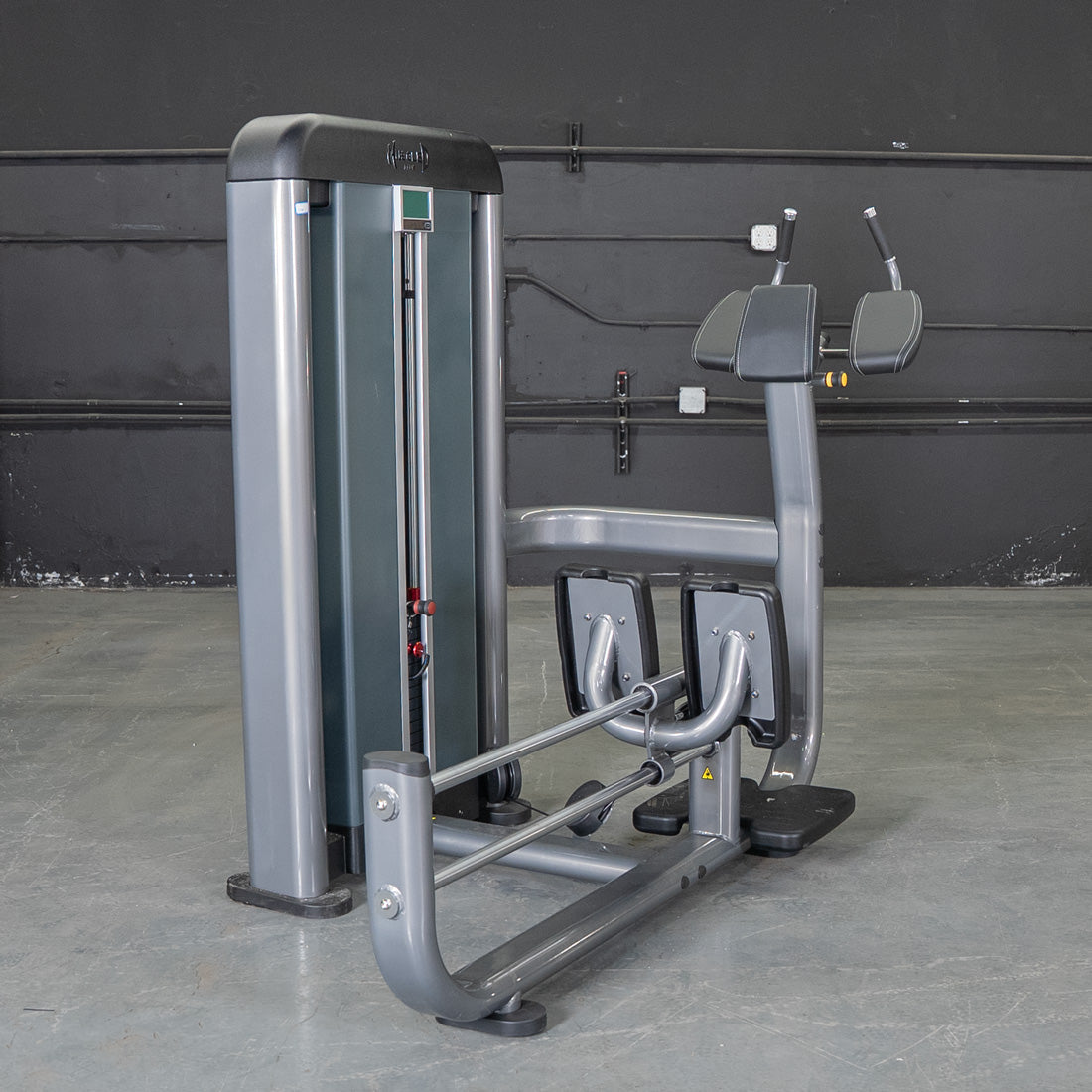 Gray commercial glute blaster machine with black padded armrests and footplates against a dark wall.