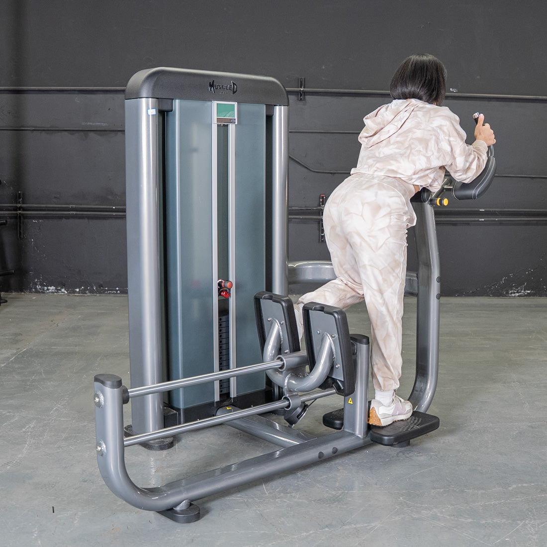 Woman using a grey glute blaster machine in a gym, pushing back with one leg.
