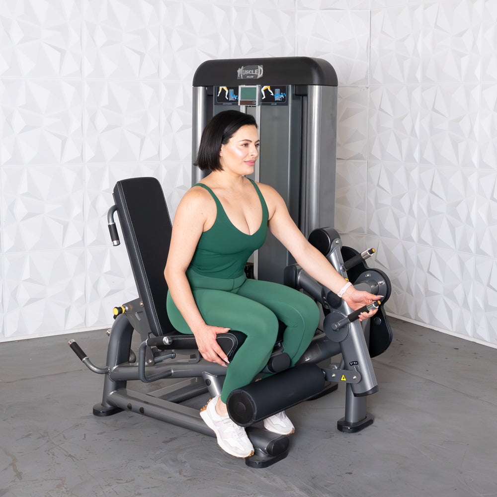 Woman in green activewear seated on a leg extension/curl machine, adjusting the settings.