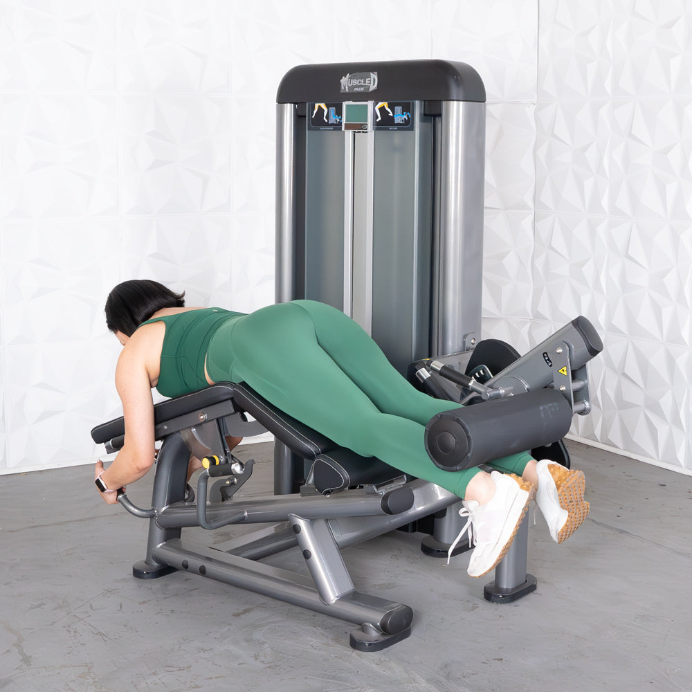 Woman in green performing a leg curl on a grey fitness machine.