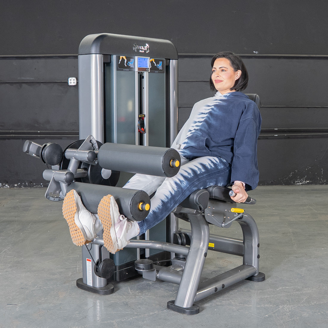Woman in tie-dye using a gray leg extension curl combo machine in a gym.