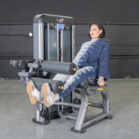 Thumbnail for Woman in tie-dye using a gray leg extension curl combo machine in a gym.