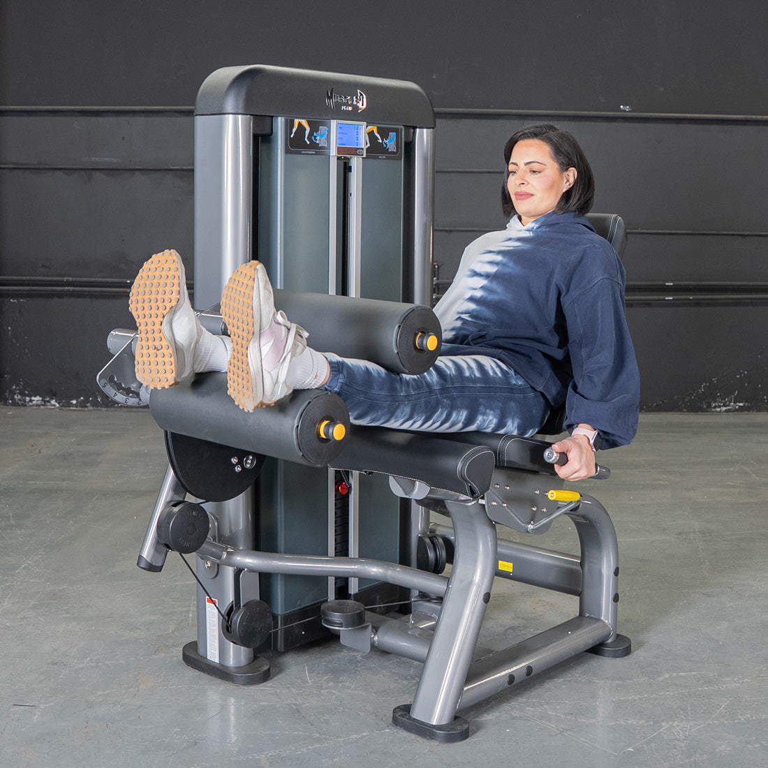 Woman using a gray and black leg extension/curl machine with yellow accents.