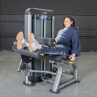 Thumbnail for Woman using a gray and black leg extension/curl machine with yellow accents.