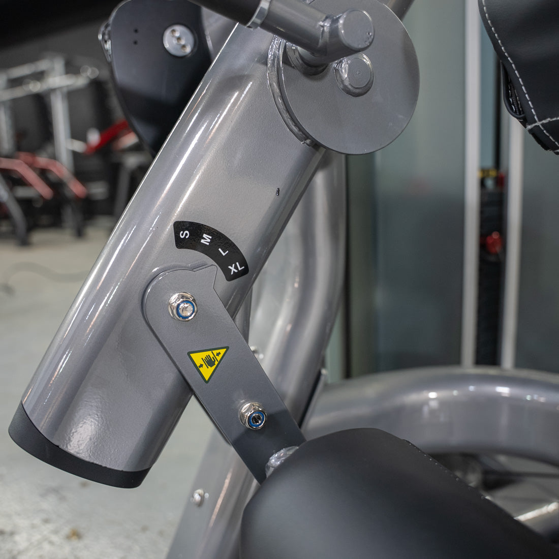 Close-up of a gray leg extension machine with S, M, L, XL size settings and a pinch point warning label.