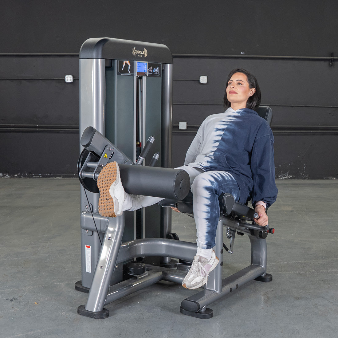 Woman using a gray leg extension machine, extending her leg with a padded bar against her shins.