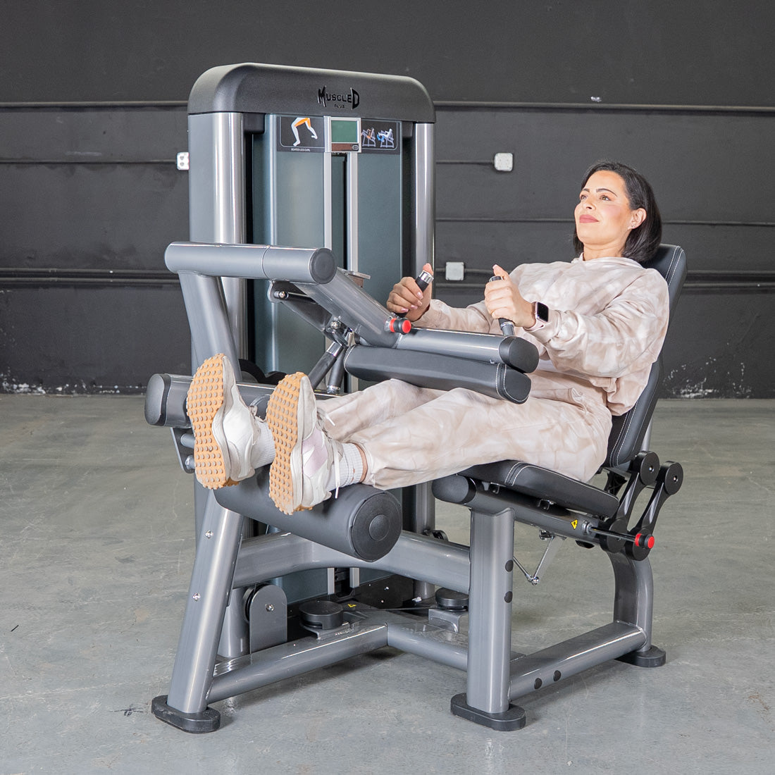 Woman in a beige sweatsuit using a seated leg curl machine in a gym.