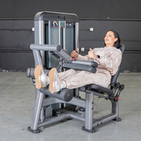 Thumbnail for Woman in a beige sweatsuit using a seated leg curl machine in a gym.