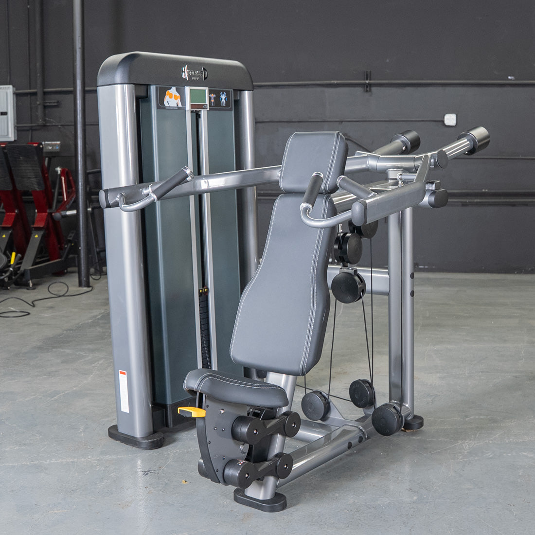 Gray shoulder press gym machine with black padded seat and handles in a gym setting.