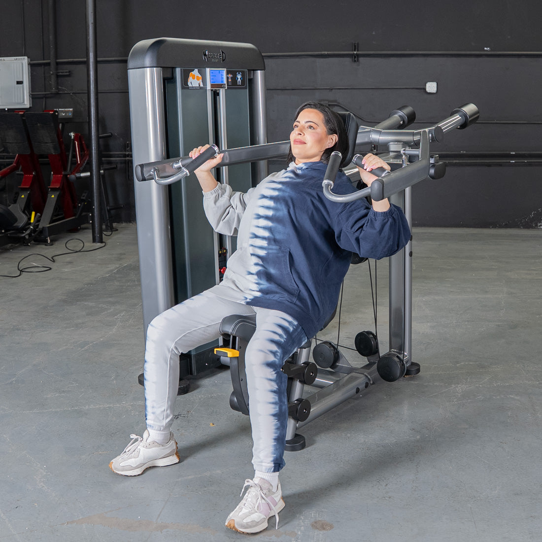 Woman in a tie-dye hoodie using an Elite Plus Shoulder Press machine at the gym.