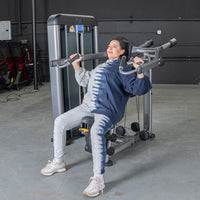 Thumbnail for Woman in a tie-dye hoodie using an Elite Plus Shoulder Press machine at the gym.