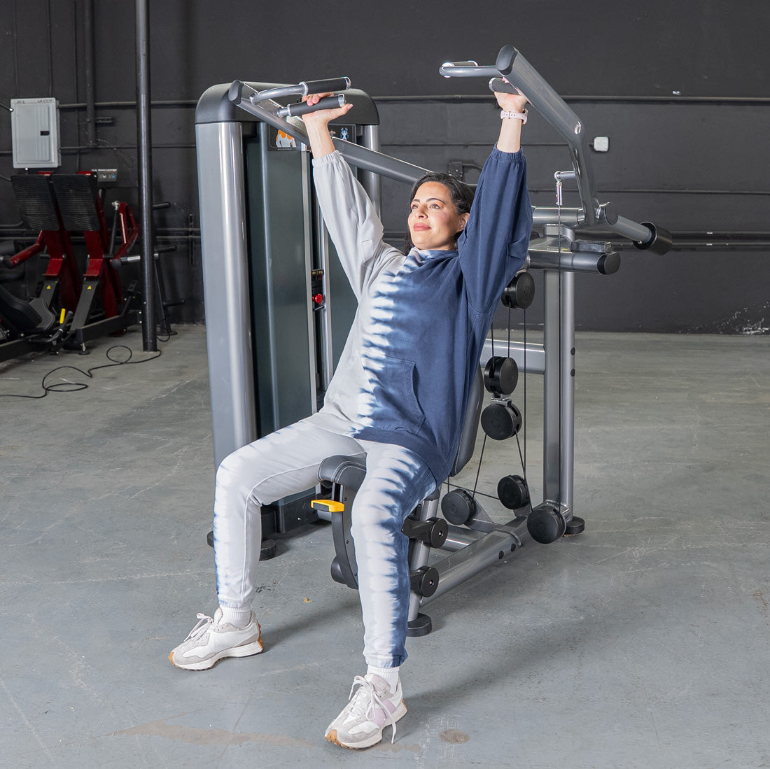 Woman in tie-dye sweats using a shoulder press machine in a gym.