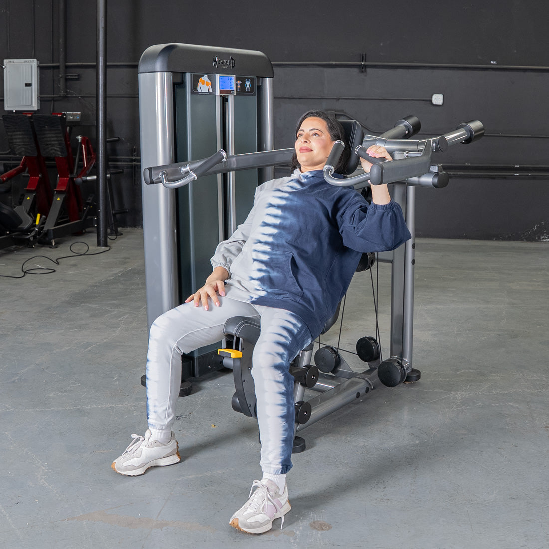 Woman in tie-dye hoodie using an isolateral shoulder press machine in a gym.