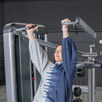 Thumbnail for Woman using a shoulder press machine, holding handles with arms extended upwards.