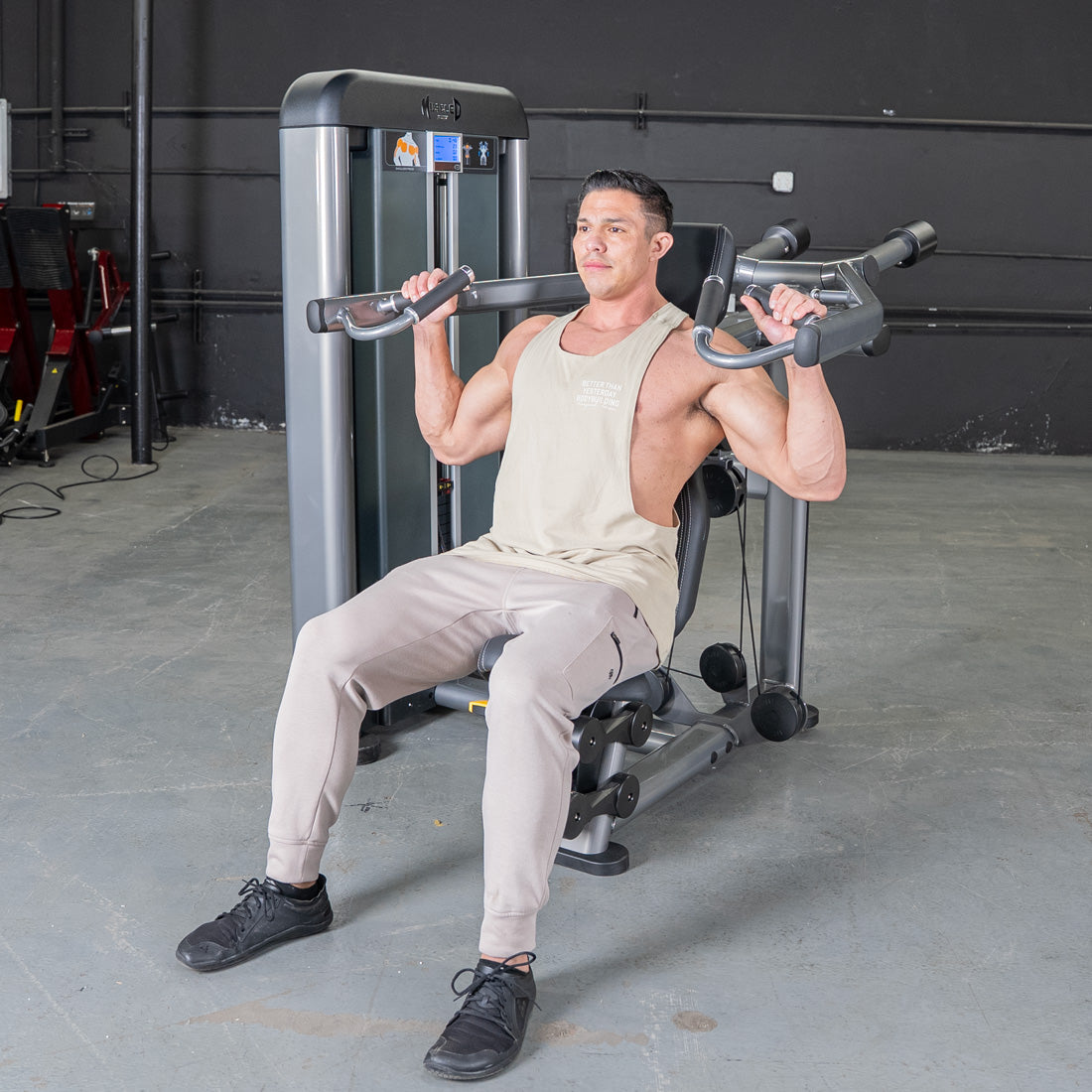 Man in a tank top and sweatpants using a shoulder press machine in a gym.