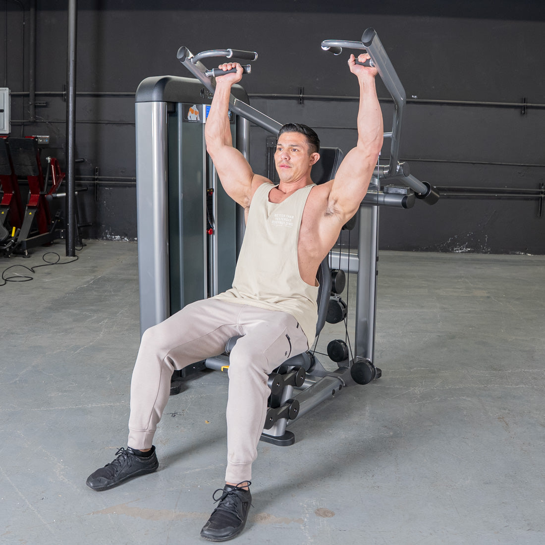 Man in a tank top and sweatpants using a shoulder press machine in a gym.