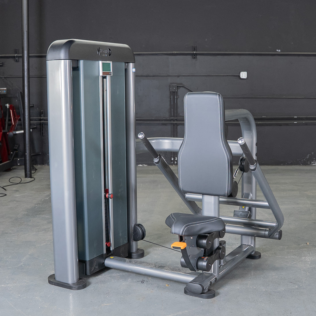 Gray triceps press machine with black padded seat and backrest, weight stack on left, in a gym setting.