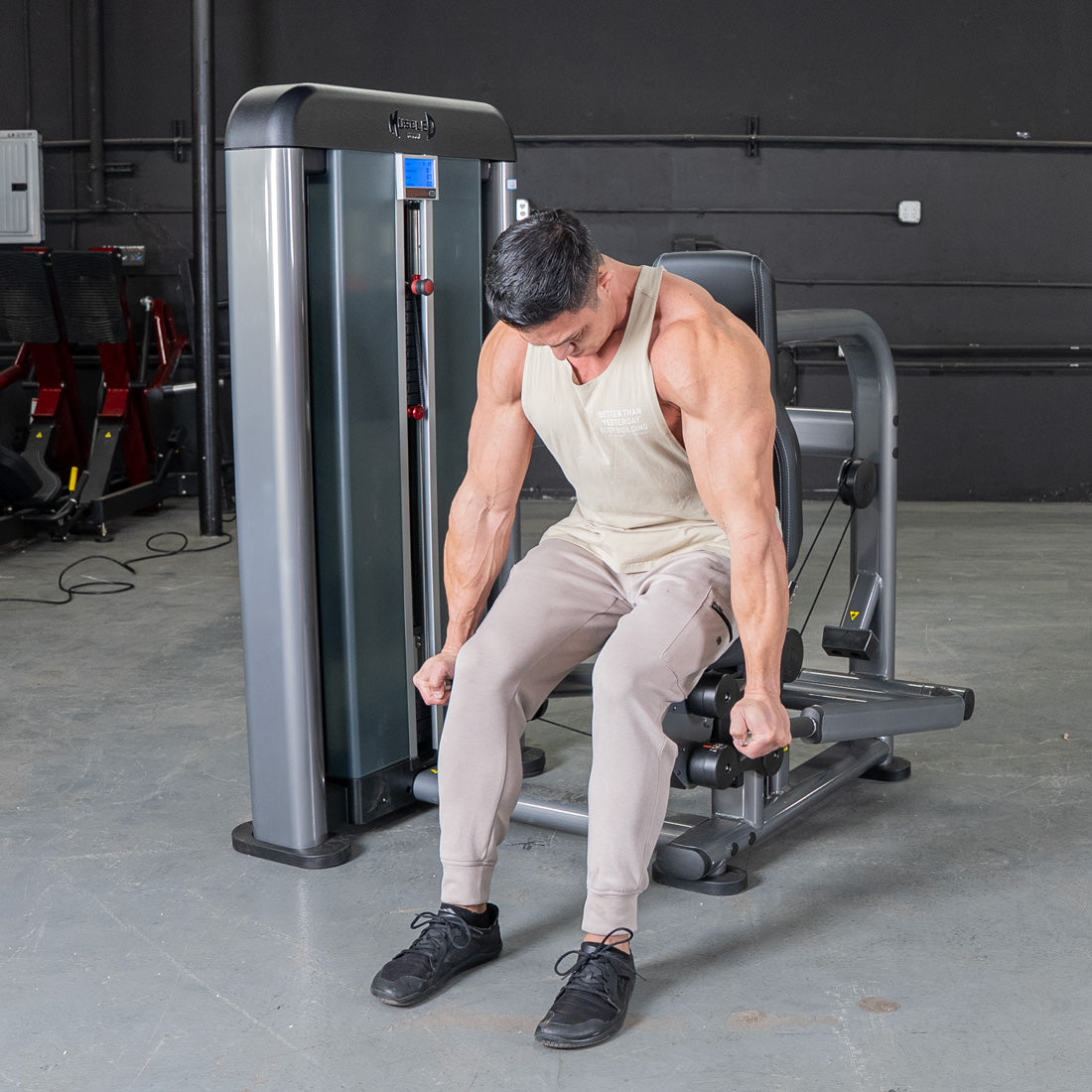 Man in a tank top and sweatpants using a triceps press machine in a gym.