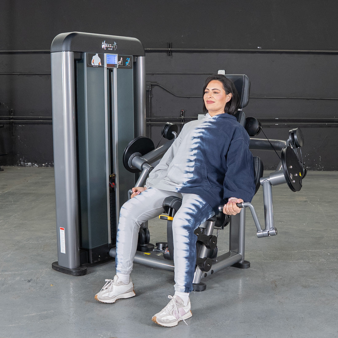 Woman in tie-dye hoodie using a gray and black biceps curl machine.