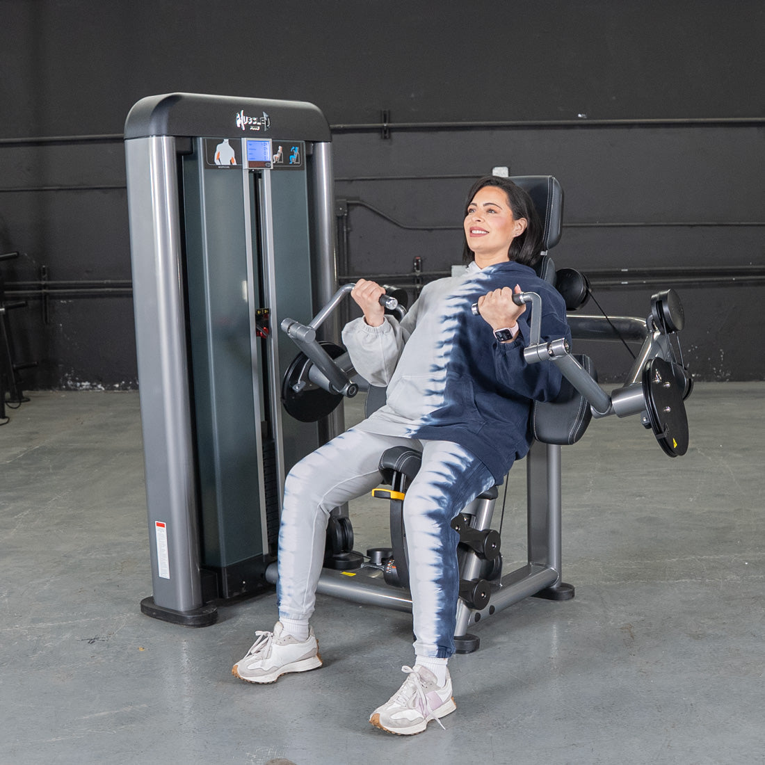 Woman using a biceps curl machine in a gym, smiling while performing the exercise.