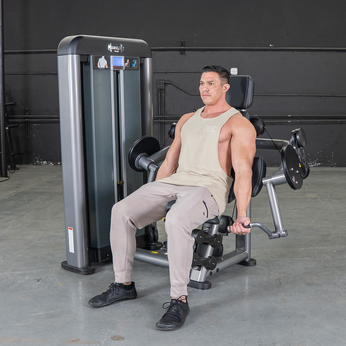 Man in a tank top and sweatpants using a gray biceps curl machine in a gym.