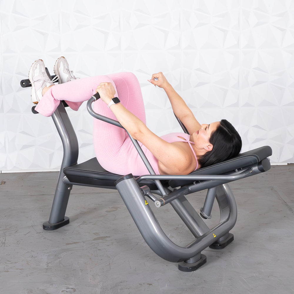 Woman in pink activewear using an ab roller machine, lying on her back with legs bent and feet secured.