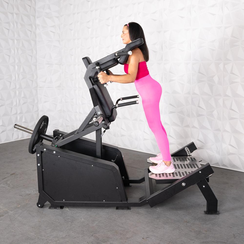 Woman in pink using a black plate-loaded squat machine with shoulder pads and a textured footplate.