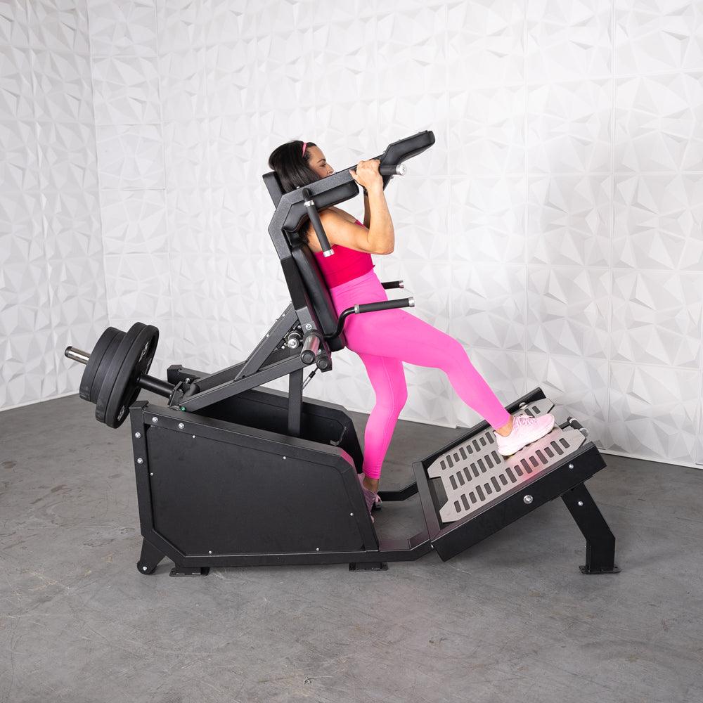 Woman in pink using a black plate-loaded hack squat machine with weights on the back.