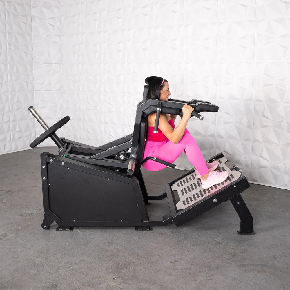 Woman in pink using a black plate-loaded hack squat machine with plate storage pegs.