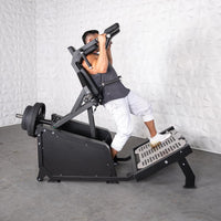 Thumbnail for Man using a black plate-loaded hack squat machine with weights on the back and a textured foot platform.