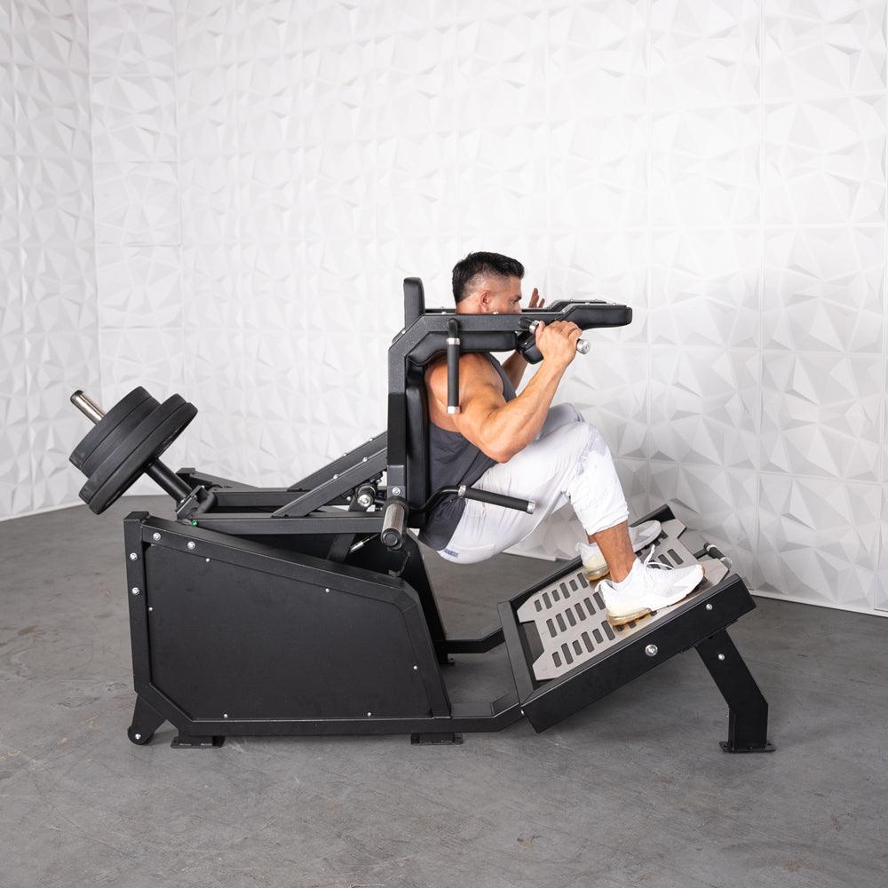 Man using a black plate-loaded hack squat machine with weights on the back and a textured white wall behind.