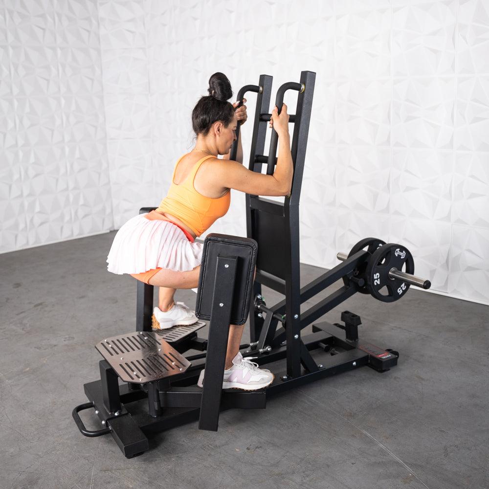 Woman using a black standing hip abductor machine with plate weights, wearing an orange top and white skirt.