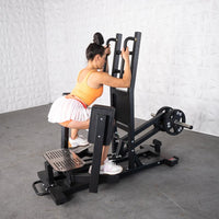 Thumbnail for Woman using a black standing hip abductor machine with plate weights, wearing an orange top and white skirt.