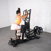 Thumbnail for Woman in orange top and white skirt using a black standing hip abductor machine with 45lb plates.