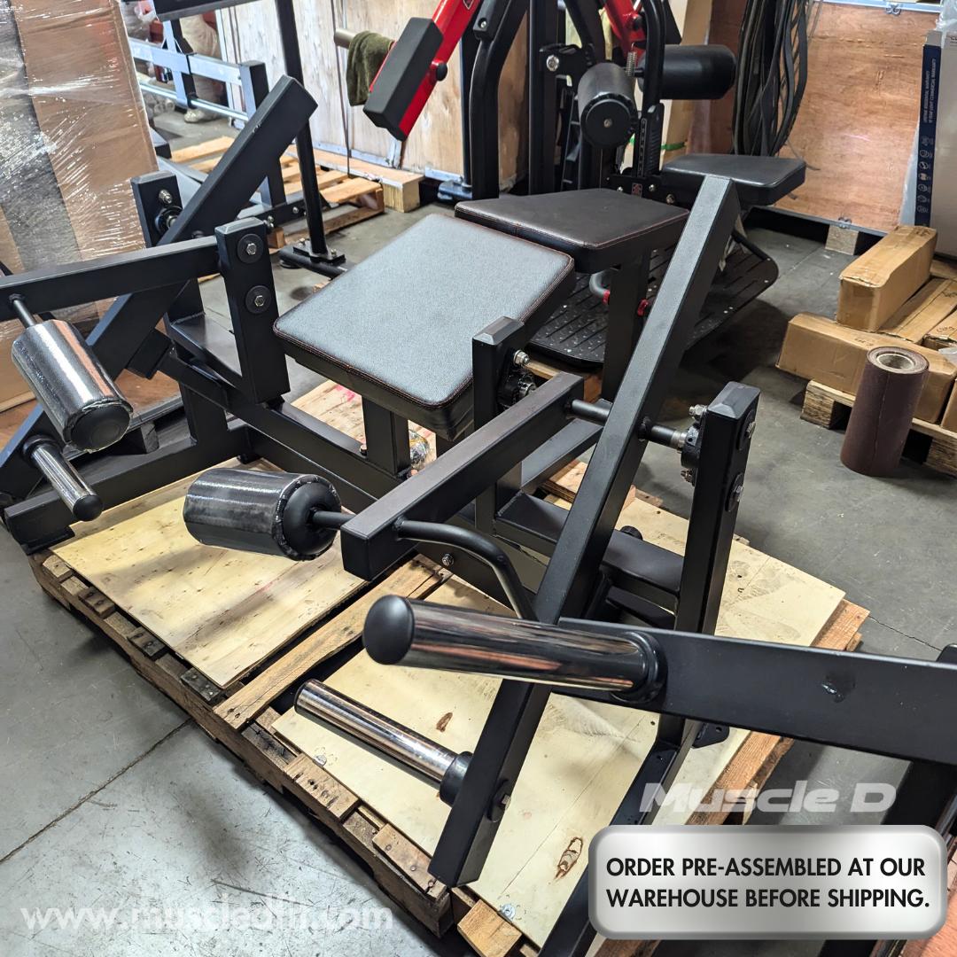 Black plate-loaded leg curl machine with gray pads on wooden pallets in a warehouse.