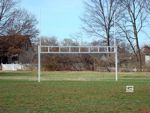 White combination soccer/football goalpost on a grassy field with trees and houses in the background.